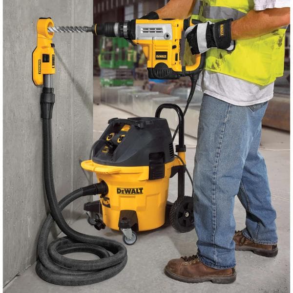 Un trabajador de la construcción utiliza un taladro DEWALT y un sistema de extracción de polvo para perforar una pared de hormigón. El equipo incluye una aspiradora DEWALT conectada por una manguera al área de perforación para minimizar el polvo durante el trabajo.