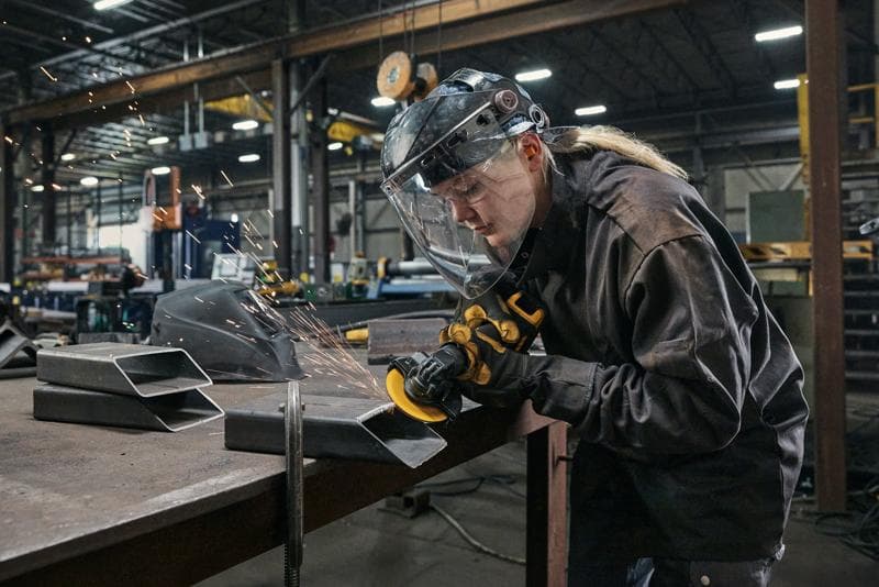 Una persona con equipo de protección utiliza una amoladora angular DEWALT DCG400 para cortar o pulir piezas de metal en un banco de trabajo dentro de un taller industrial. Se pueden ver chispas y el entorno contiene varias herramientas y equipos.