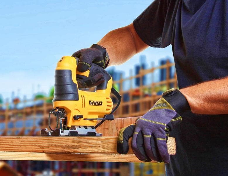 Una persona con guantes de protección está usando una sierra de calar amarilla DEWALT para cortar un trozo de madera, con una obra en construcción al fondo.