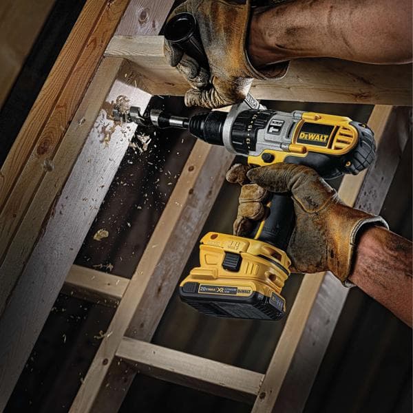 Una persona que lleva guantes de trabajo marrones utiliza un taladro inalámbrico DEWALT para perforar un agujero en una viga de madera, con virutas de madera volando durante el proceso.