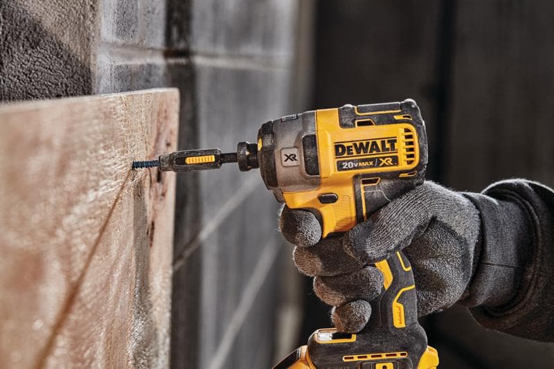 Una persona con guantes de trabajo utiliza un destornillador de impacto inalámbrico DEWALT 20V MAX XR amarillo y negro para atornillar un tornillo en una tabla de madera.