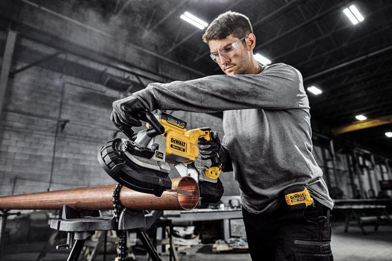 Una persona utiliza una sierra de banda portátil DEWALT DCS374B para cortar una tubería de metal en un taller industrial. La cara de la persona está desenfocada por privacidad.