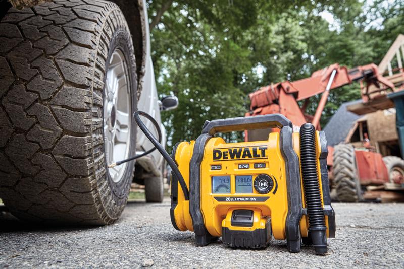 Se muestra un inflador de neumáticos portátil DEWALT en uso, conectado a la llanta de un vehículo, en un sitio de trabajo al aire libre con equipo de construcción al fondo.